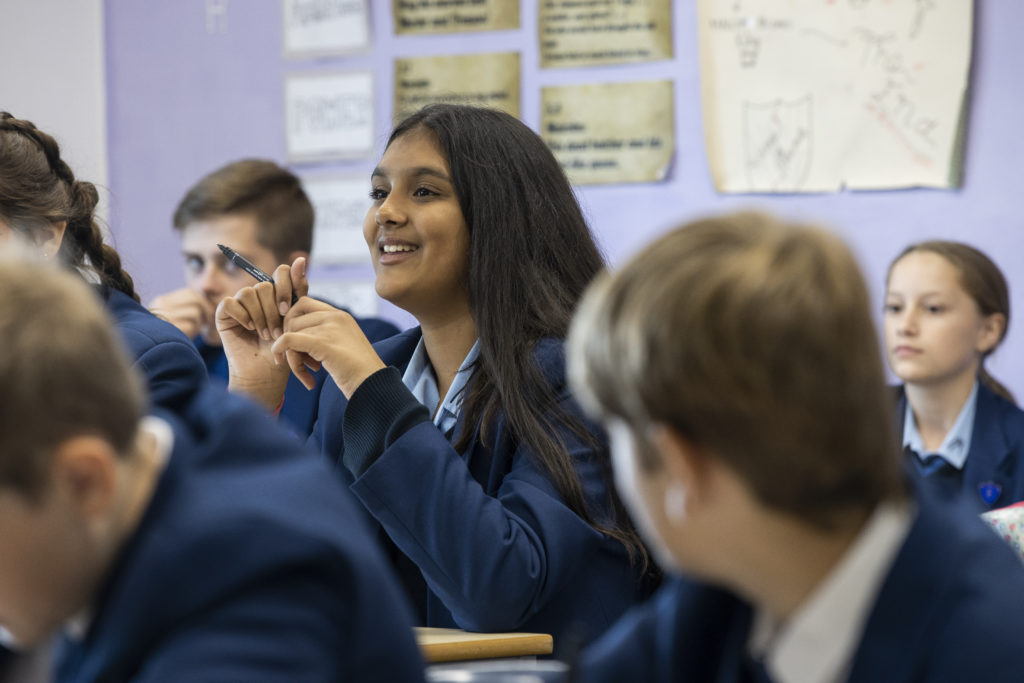 A female student is seen smiling at her desk, with her eyes looking towards the front of the class.